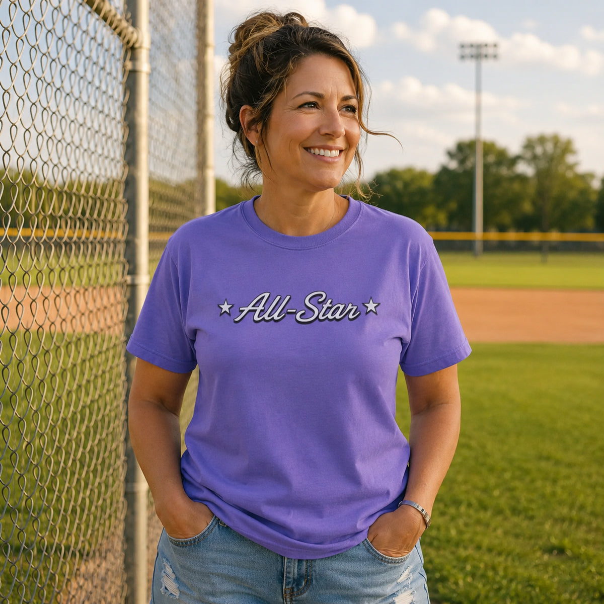 Woman wearing The Rad Rewind All-Star Graphic Tee in Violet at a baseball diamond, chain-link fence and grass behind her