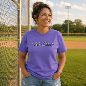 Woman wearing The Rad Rewind All-Star Graphic Tee in Violet at a baseball diamond, chain-link fence and grass behind her