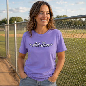 Woman wearing The Rad Rewind All-Star Graphic Tee in Violet at a baseball diamond, chain-link fence and outfield grass behind her