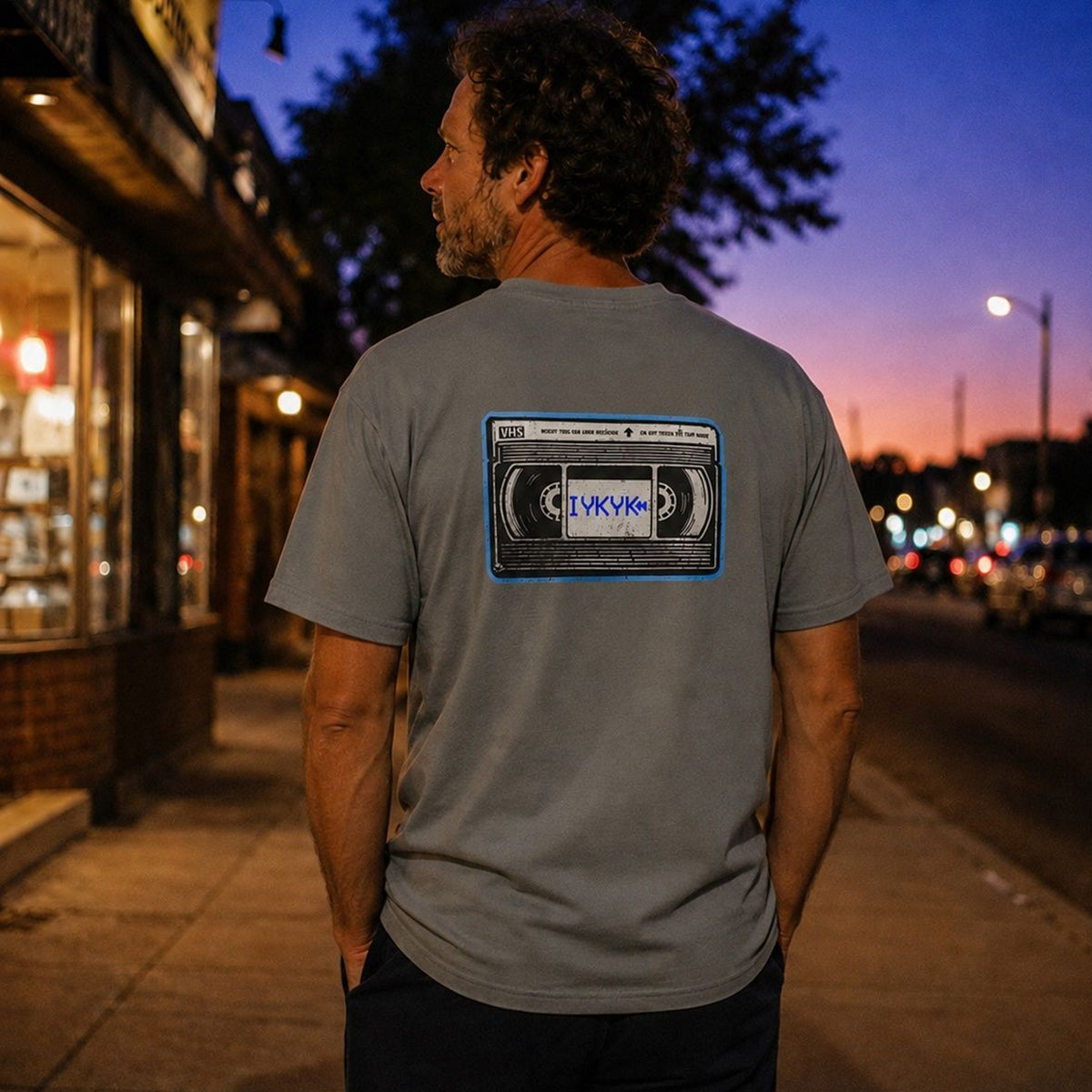 The Rad Rewind IYKYK Graphic Tee in Granite — lifestyle back shot at twilight showing VHS tape graphic
