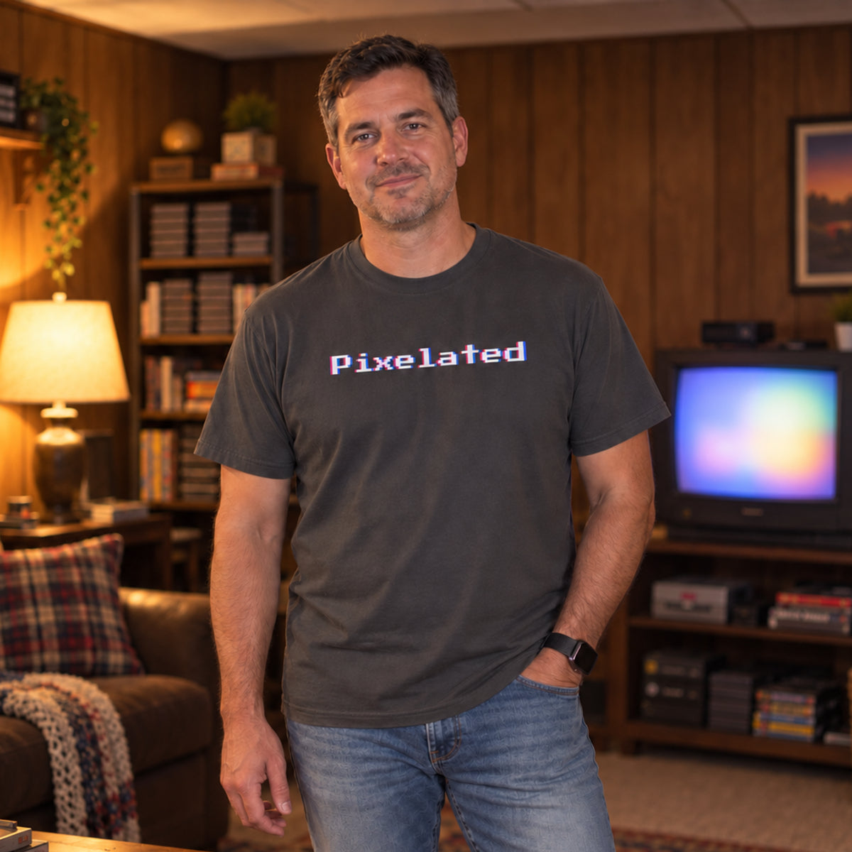 Man in his forties wearing the Graphite pixelated glitch tee in a wood-paneled rec room, showing the 8-bit wordmark and relaxed fit.
