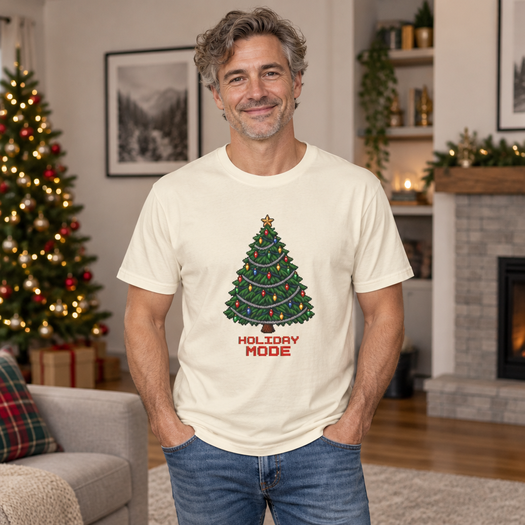 Smiling man with gray hair wearing an Ivory cream Holiday Mode pixel Christmas tree tee in a living room with a fireplace