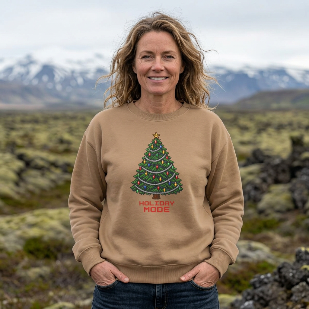 Woman with wavy hair wearing a Sandstone tan Holiday Mode pixel Christmas tree sweatshirt outdoors with snowcapped mountains behind her