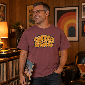 Laughing man in glasses wearing the Brick Outta Sight graphic tee in a wood-paneled rec room with a turntable