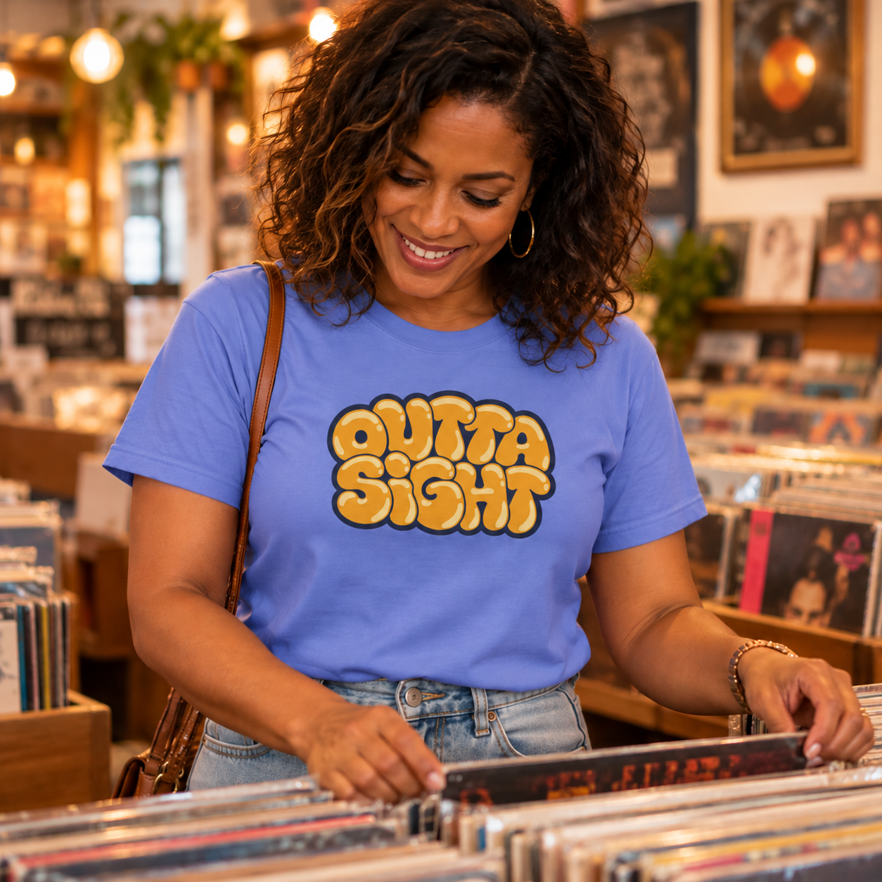 Smiling woman with curly hair wearing the Flo Blue Outta Sight graphic tee while flipping through records in a record store