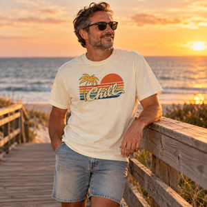 Man in sunglasses leaning on a beach boardwalk railing at sunset, wearing the Ivory Chill graphic tee