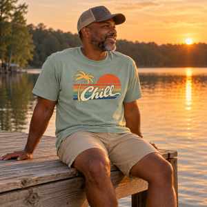 Man in a trucker cap sitting on a lake dock at sunset, wearing the Bay Chill graphic tee