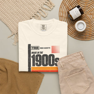 Ivory Made in the 1900s retro cassette label graphic tee folded in a flat lay with chinos, a woven basket, and a knit throw