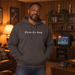 Man in his forties wearing the Charcoal pixelated glitch hoodie in a wood-paneled rec room with a tube TV, showing the 8-bit chest wordmark and relaxed fit.