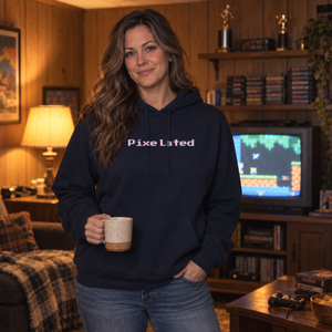 Woman in her late thirties wearing the Navy pixelated glitch hoodie in a wood-paneled rec room, holding a mug beside a tube TV, showing the 8-bit chest wordmark and relaxed fit.