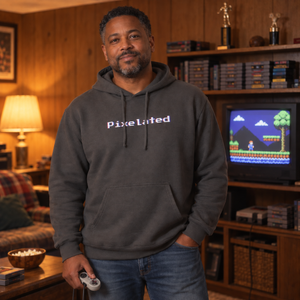 Man in his forties wearing the Charcoal pixelated glitch hoodie in a wood-paneled rec room with a tube TV, showing the 8-bit chest wordmark and relaxed fit.