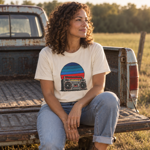 Retro boombox sunset tee in ivory worn by a woman seated on a weathered pickup tailgate — The Rad Rewind